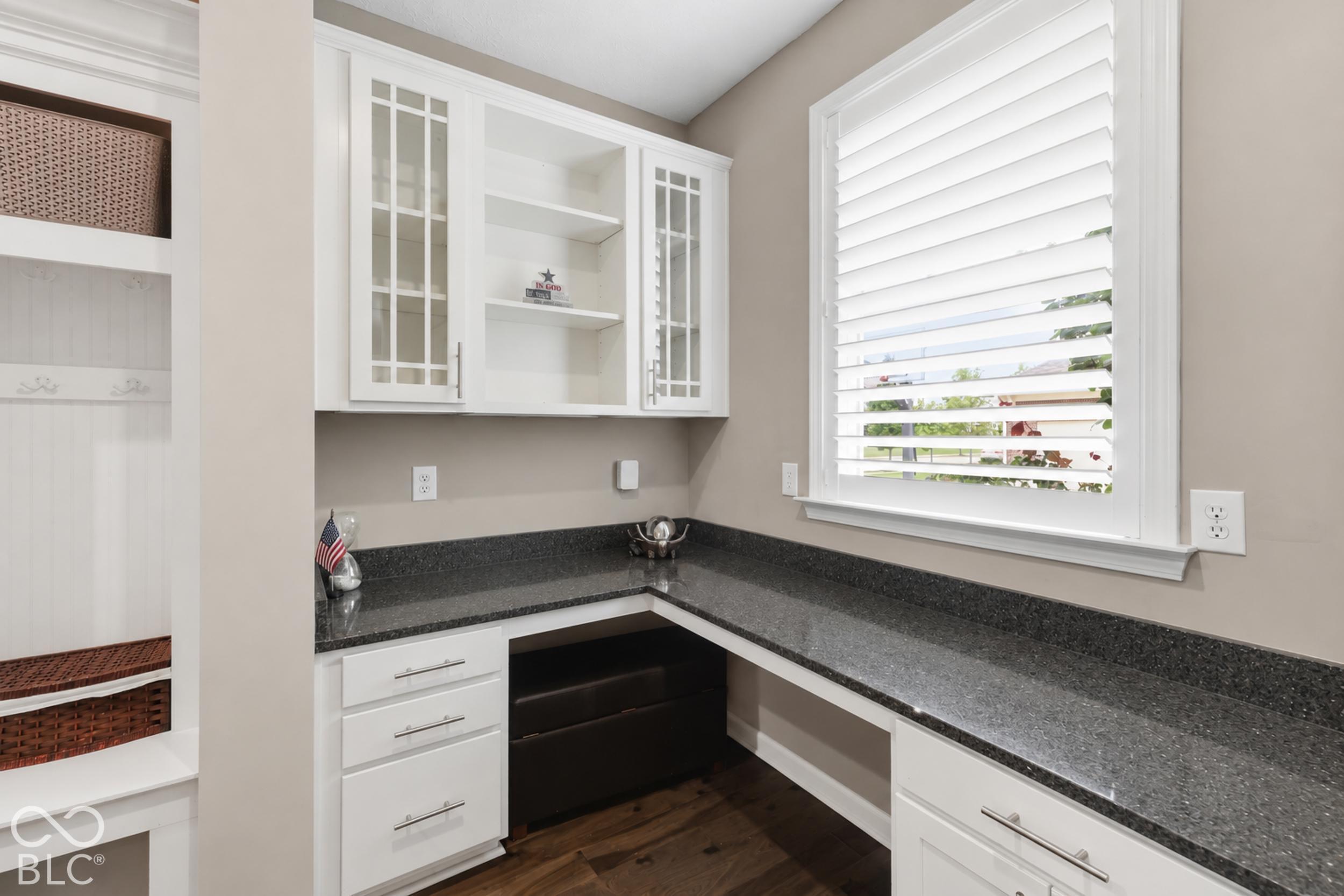 Home office with dark L-shaped countertops, white cabinetry, built-in basket shelves, and a shuttered window.