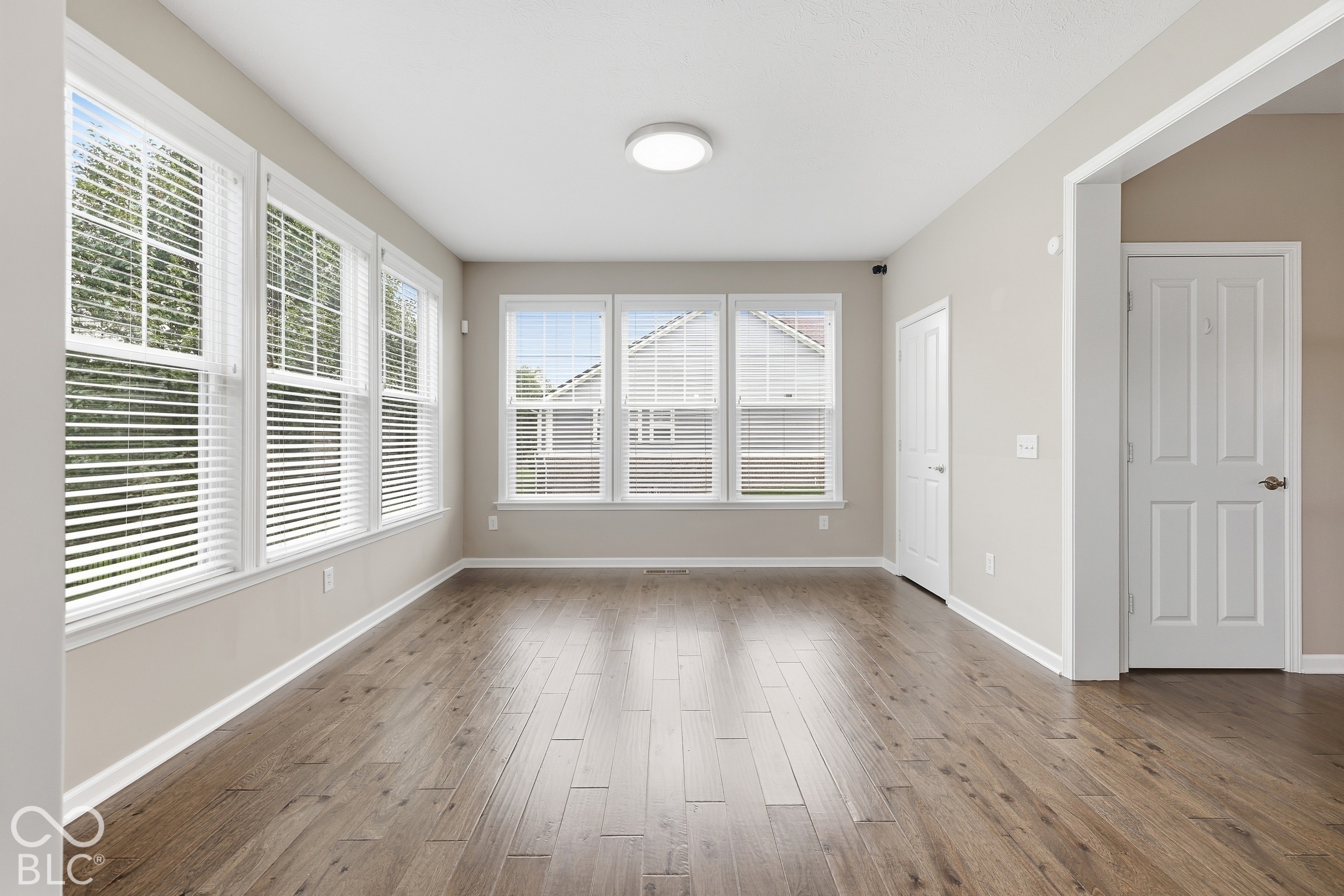 Empty room with wood flooring and large windows fitted with white blinds.