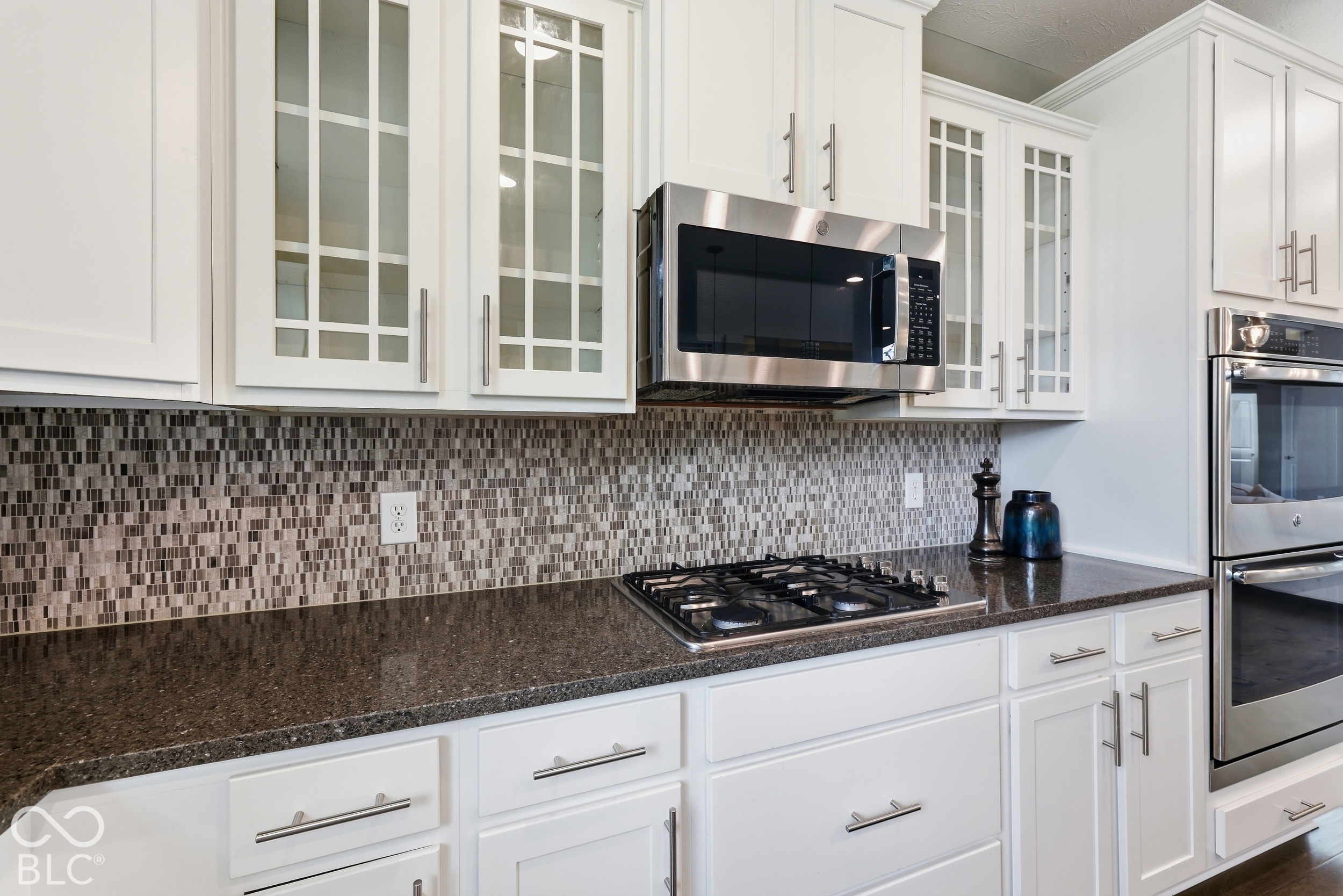 Kitchen with white glass-front cabinets, mosaic backsplash, granite counters, and stainless steel appliances.