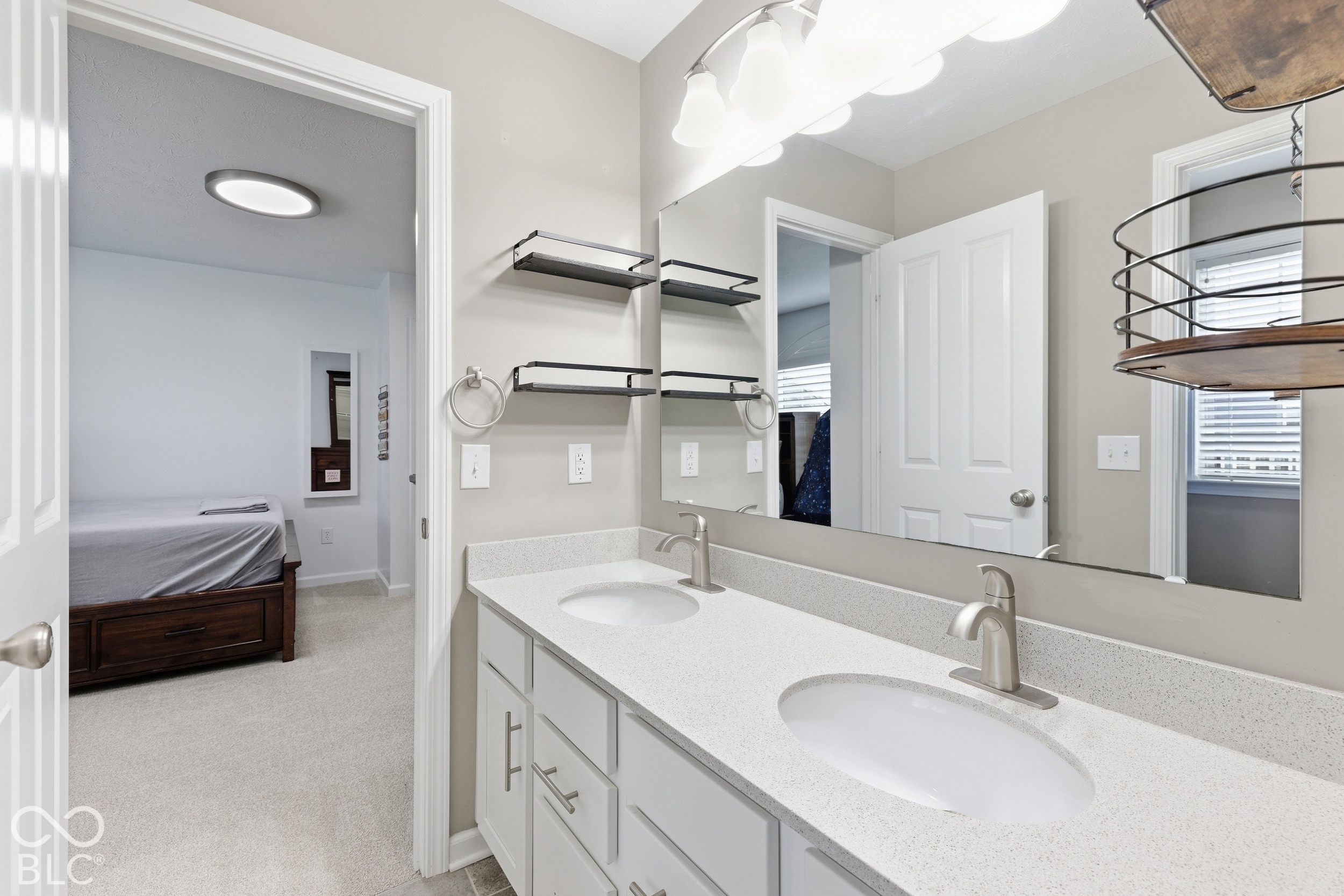 Bathroom with a long double-sink vanity and mirror beside an open doorway to a bedroom.