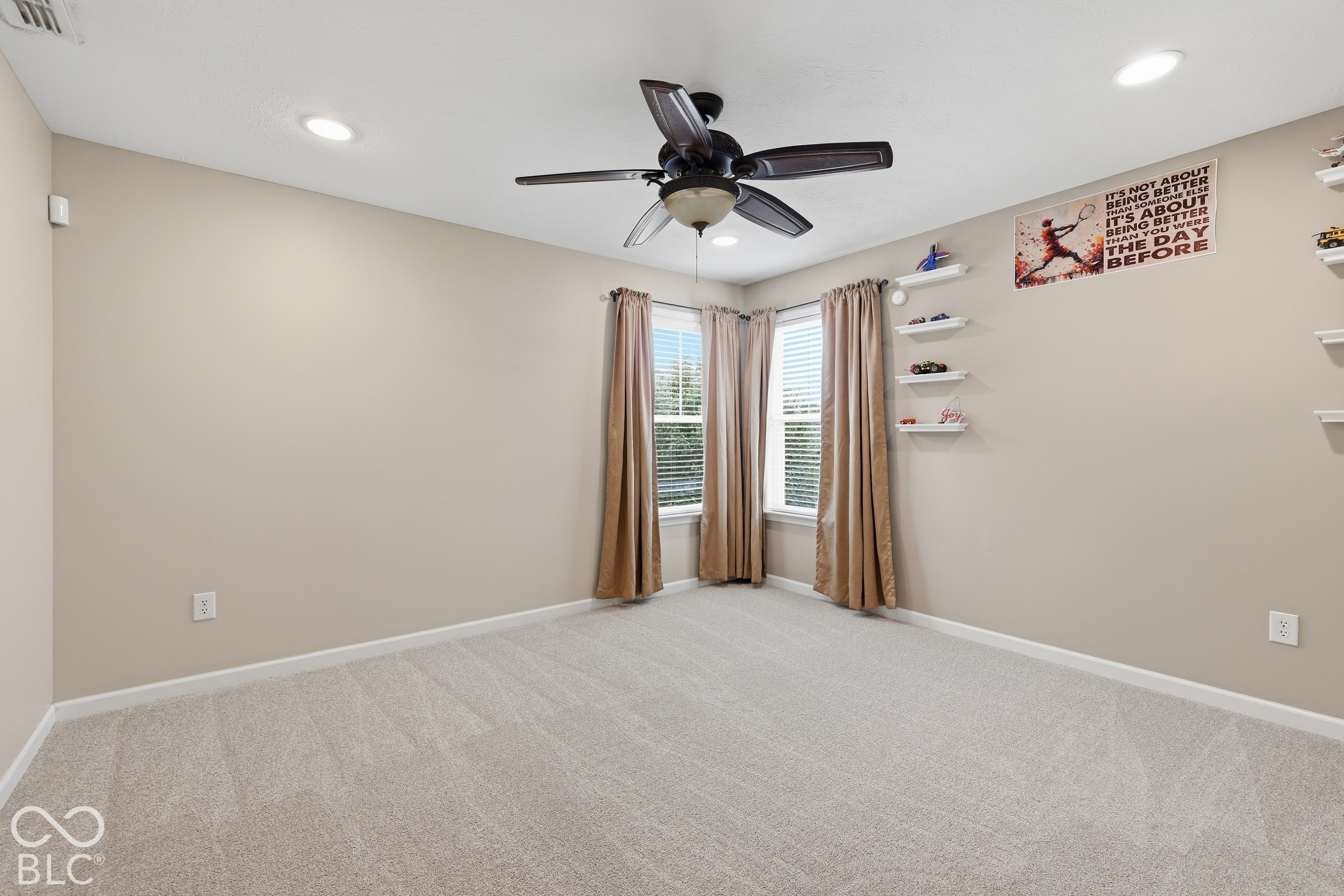 Empty carpeted bedroom with a dark ceiling fan, corner windows, tan curtains, and white display shelves.