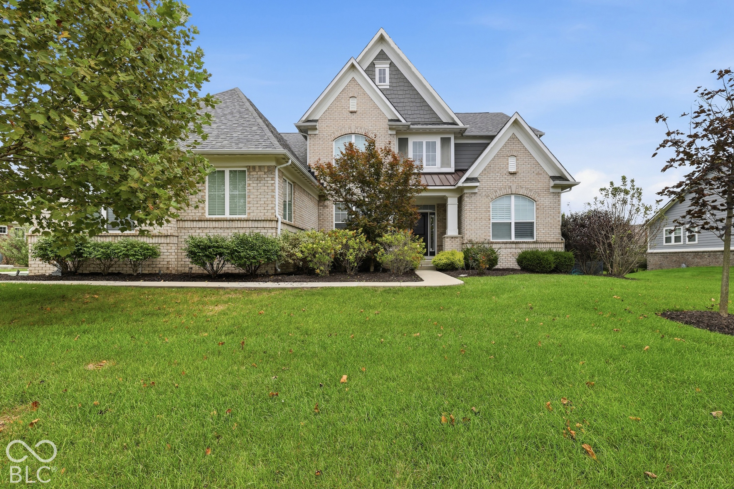 Two-story beige brick home with steep gables, front shrubs, and a broad green lawn.