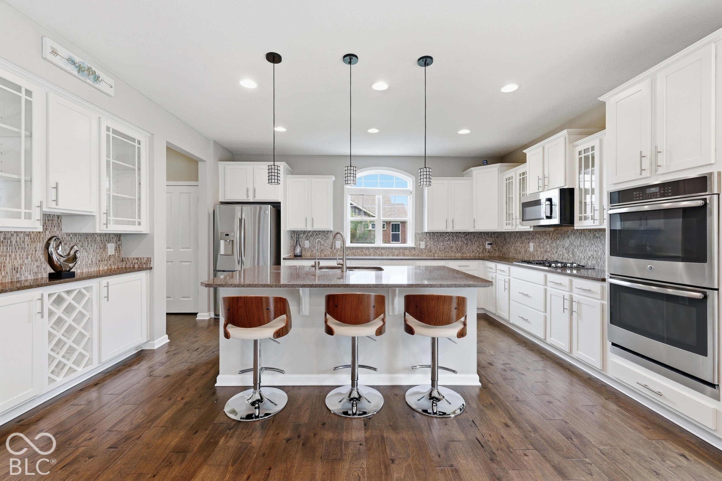 Bright white kitchen with a large island, three bar stools, stainless steel appliances, and dark wood floors.