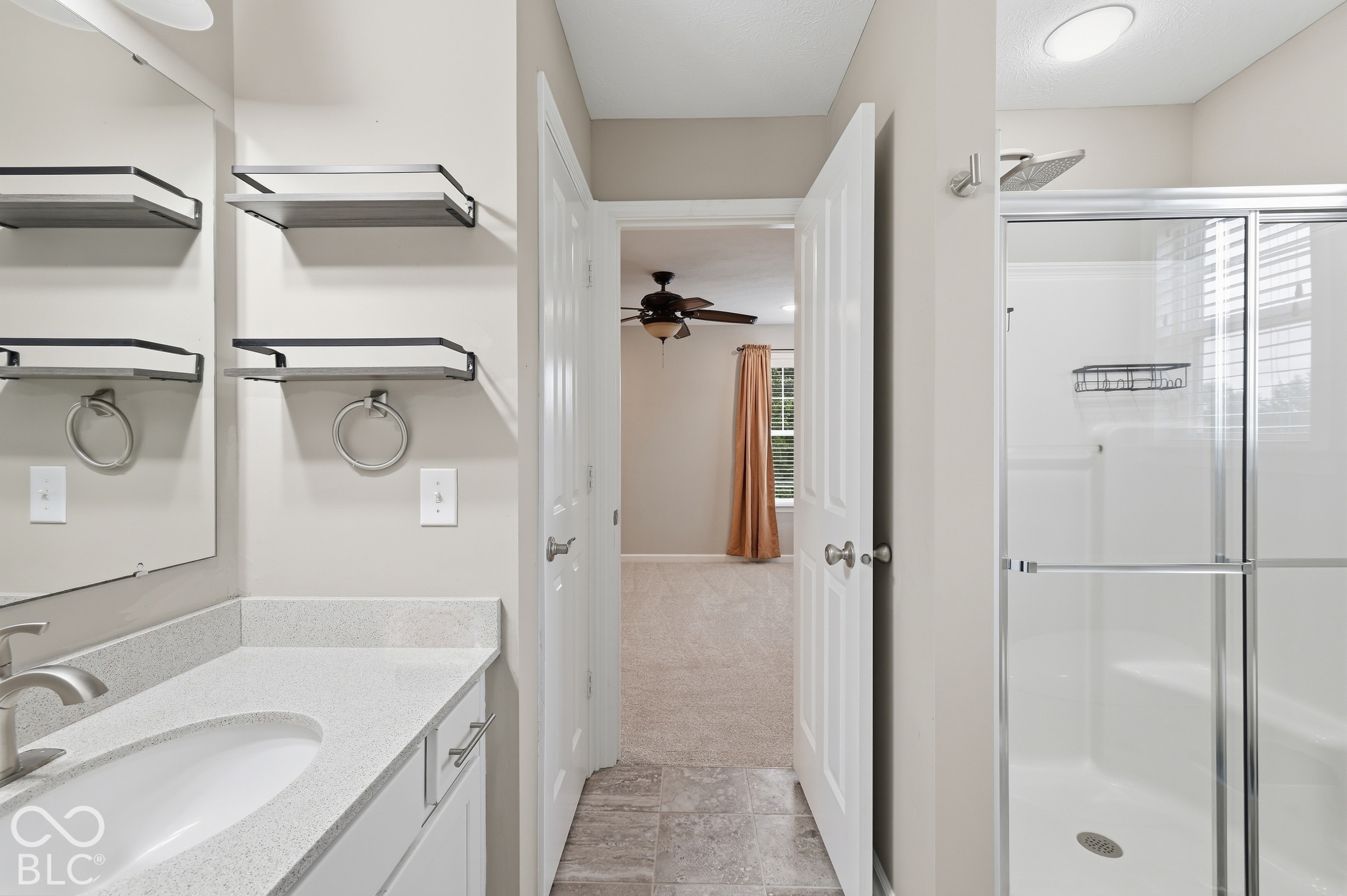 Bathroom with a glass-enclosed shower and white vanity, looking through an open doorway into a carpeted bedroom.