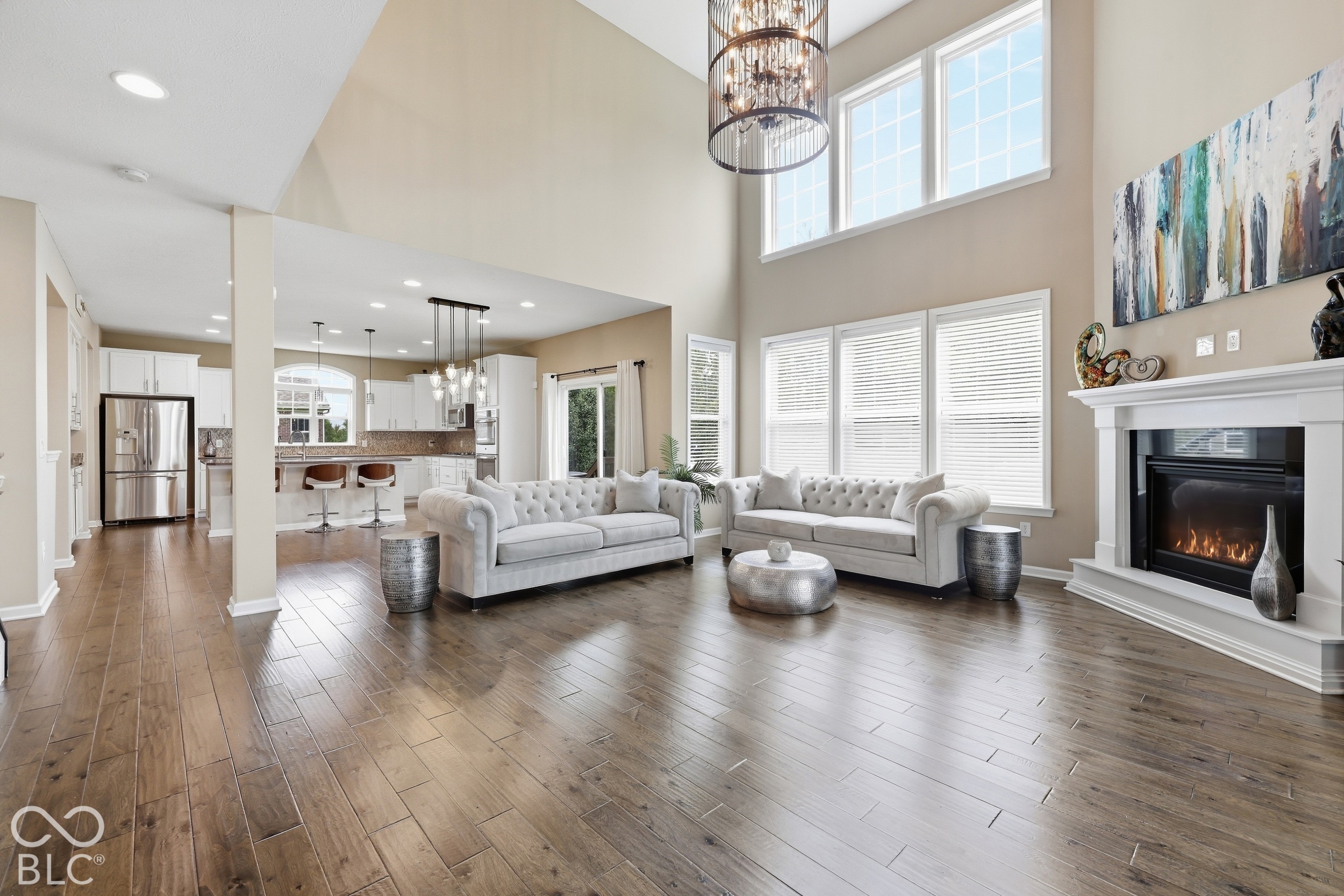 Living room with tall windows, white sofas, a fireplace, and wood floors, open to the kitchen.