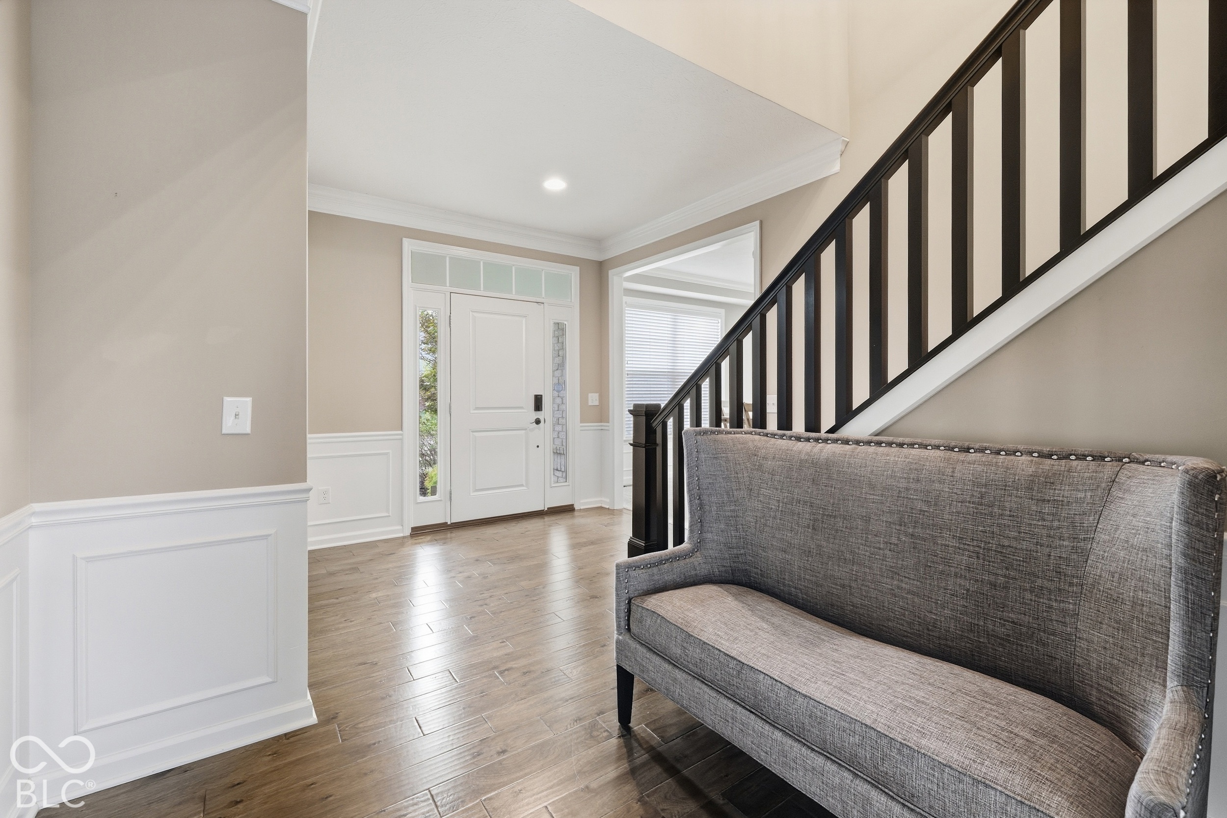 Foyer with gray upholstered bench, dark staircase, white entry door, and wood-look flooring.