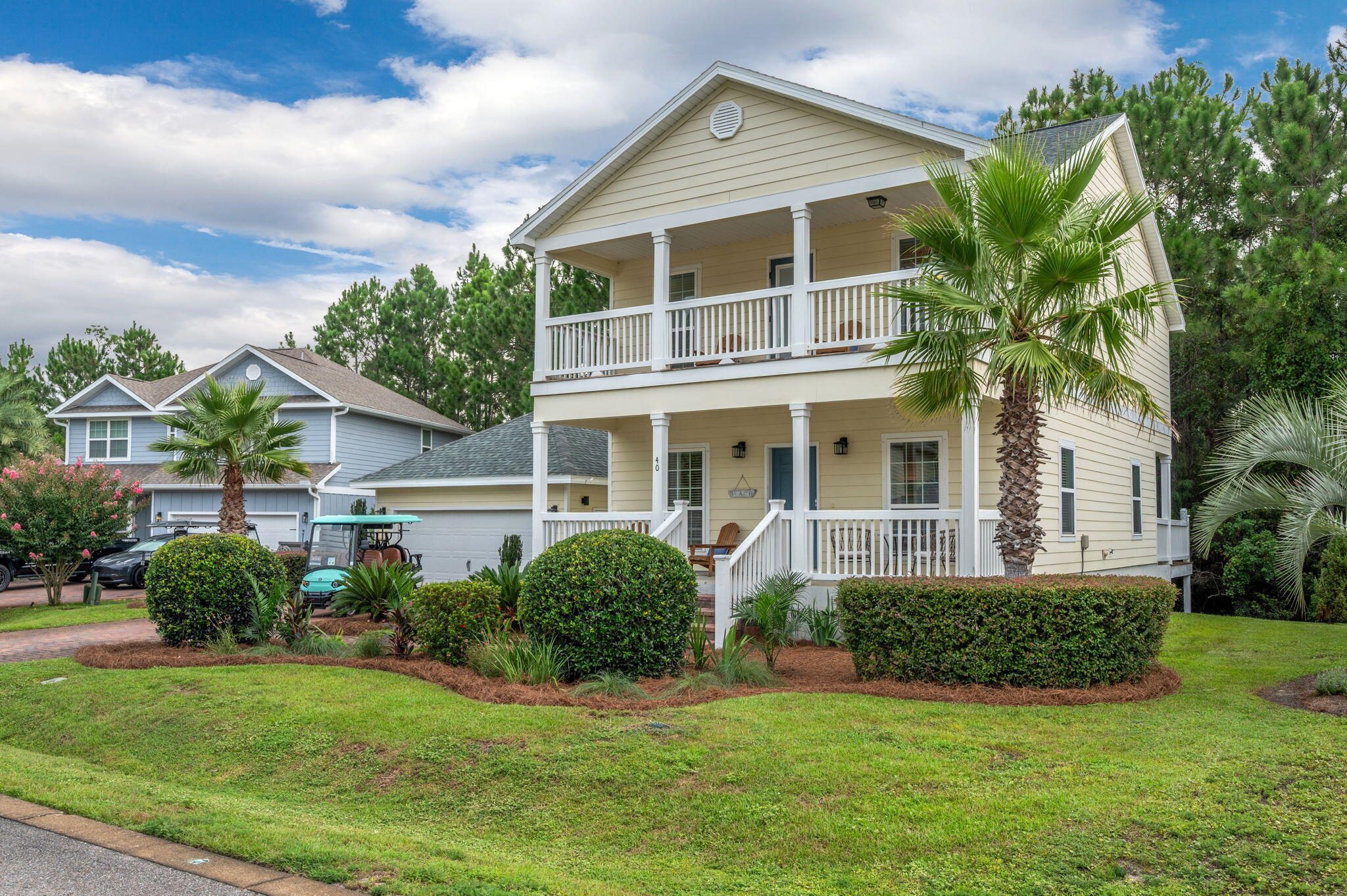 Two-story beige home with white wraparound porches, palm trees, and a landscaped front lawn.