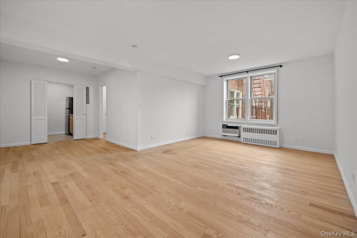 Vacant living room with hardwood floors, recessed lights, and a window overlooking a neighboring brick building.