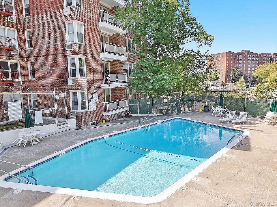 Outdoor swimming pool beside a brick apartment building, with lounge chairs and mature trees.
