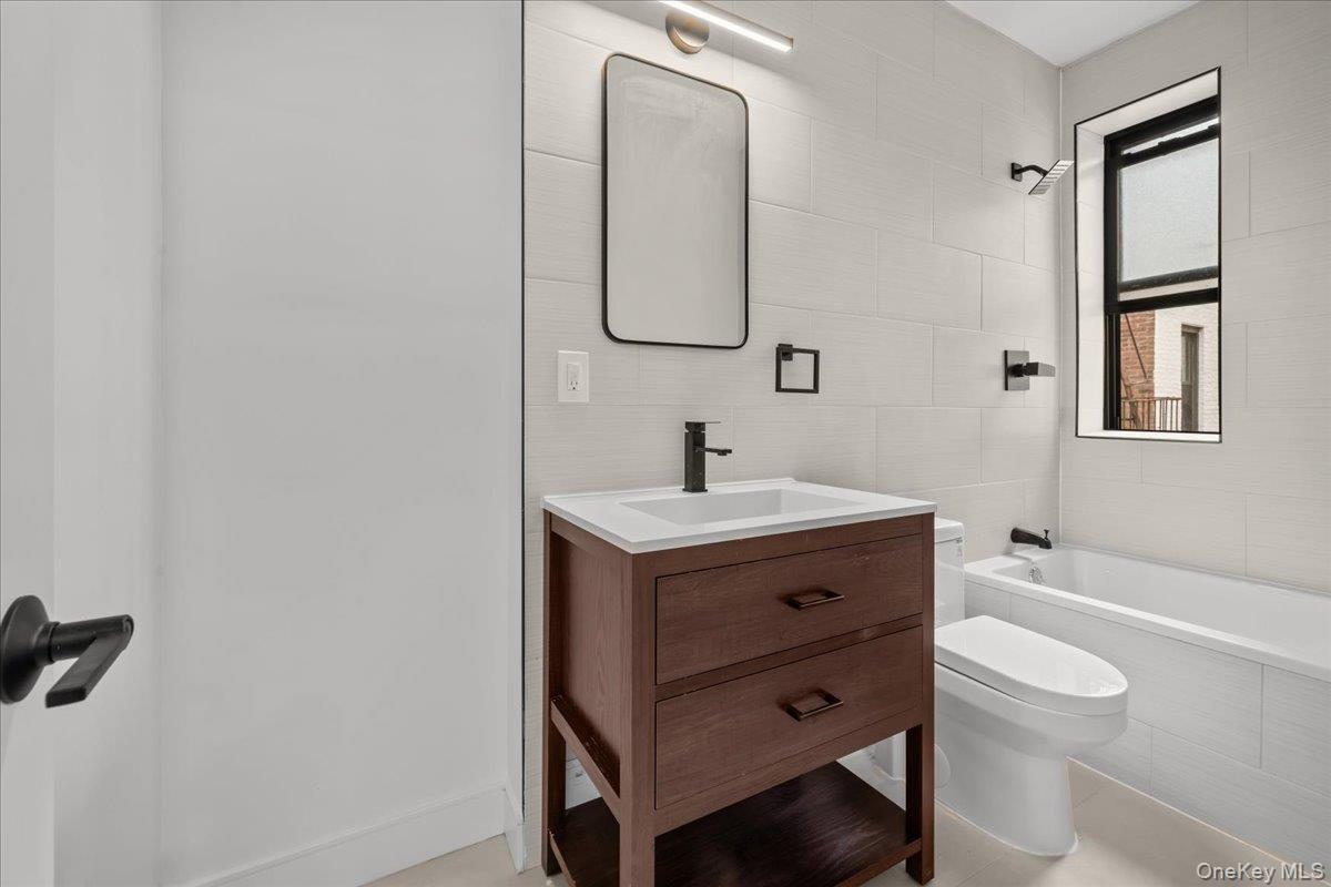 Renovated bathroom with a dark wood vanity, white tiled walls, and a bathtub beside a black-framed window.