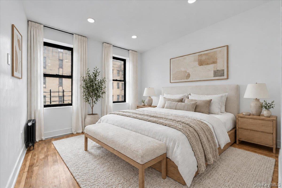 Staged bedroom with a king-size bed, upholstered bench, hardwood floors, and two curtained windows.