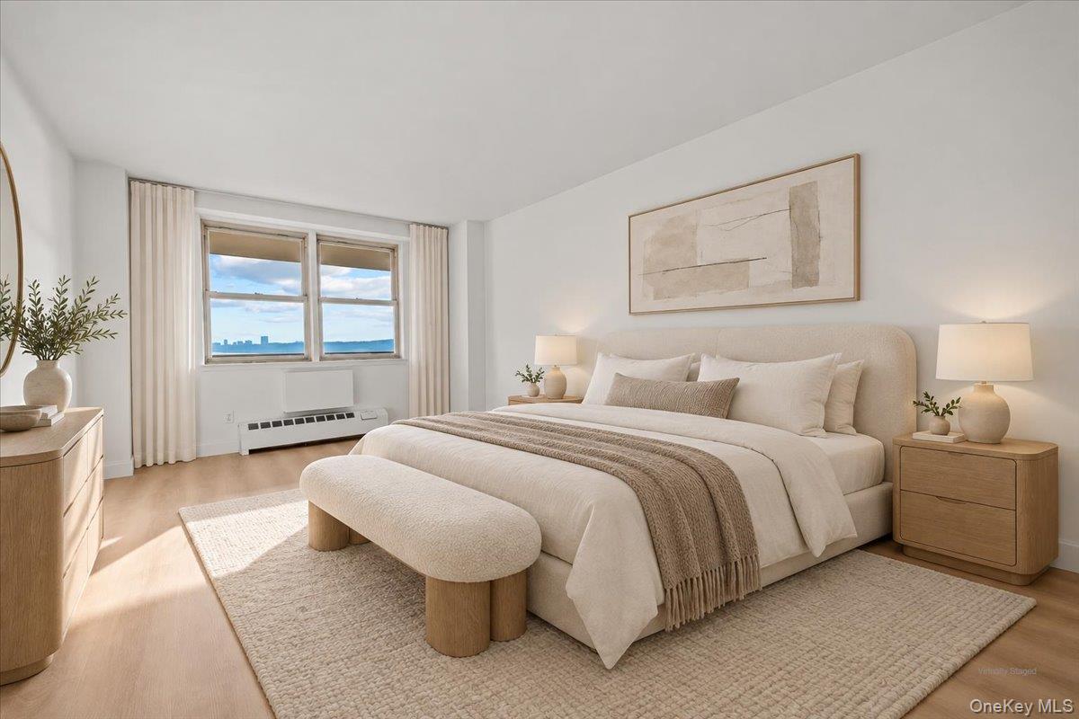 Staged bedroom with a large neutral bed, cushioned bench, and windows overlooking water.