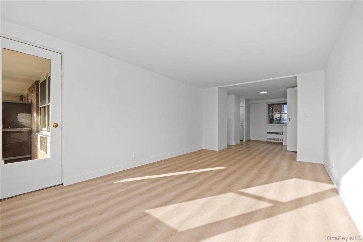 Vacant living room with light wood floors, white walls, and sunlight streaming across the floor.