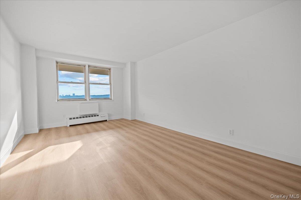 Vacant room with light wood floors, two windows, and distant water and skyline views.