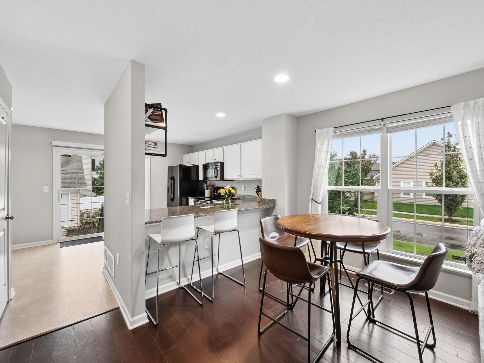 Open kitchen and dining area with a breakfast bar, white stools, and a round table by large windows.