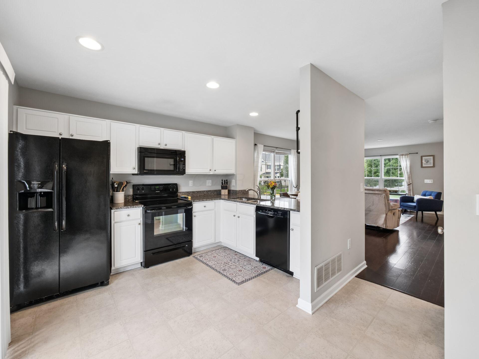 Kitchen with white cabinets and black appliances opening into a living room with large windows.