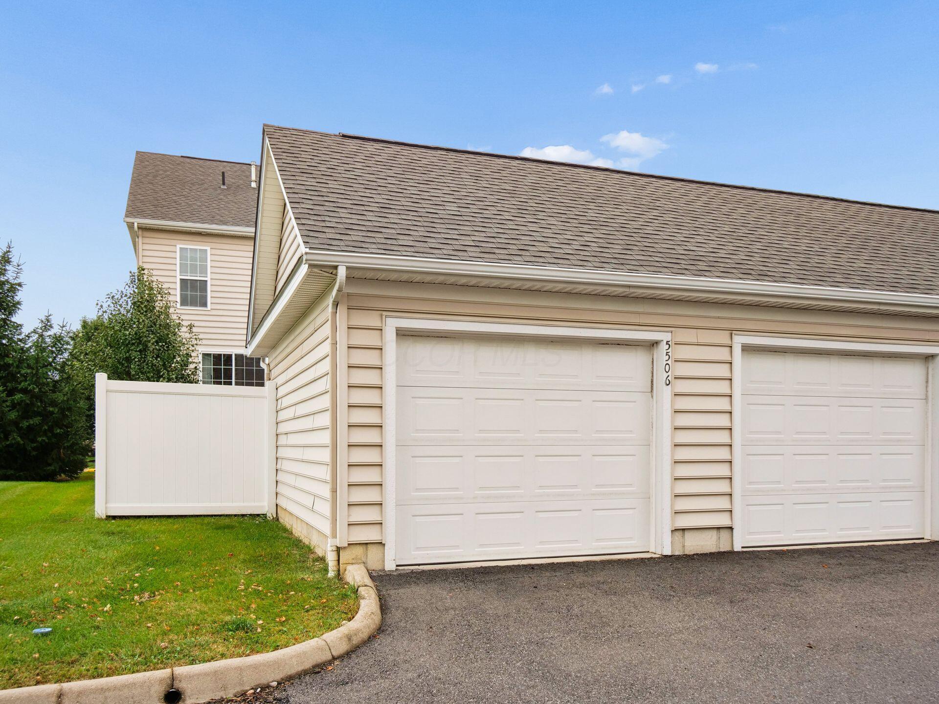 Beige-sided property with two white garage doors, a white fence, and a green lawn.