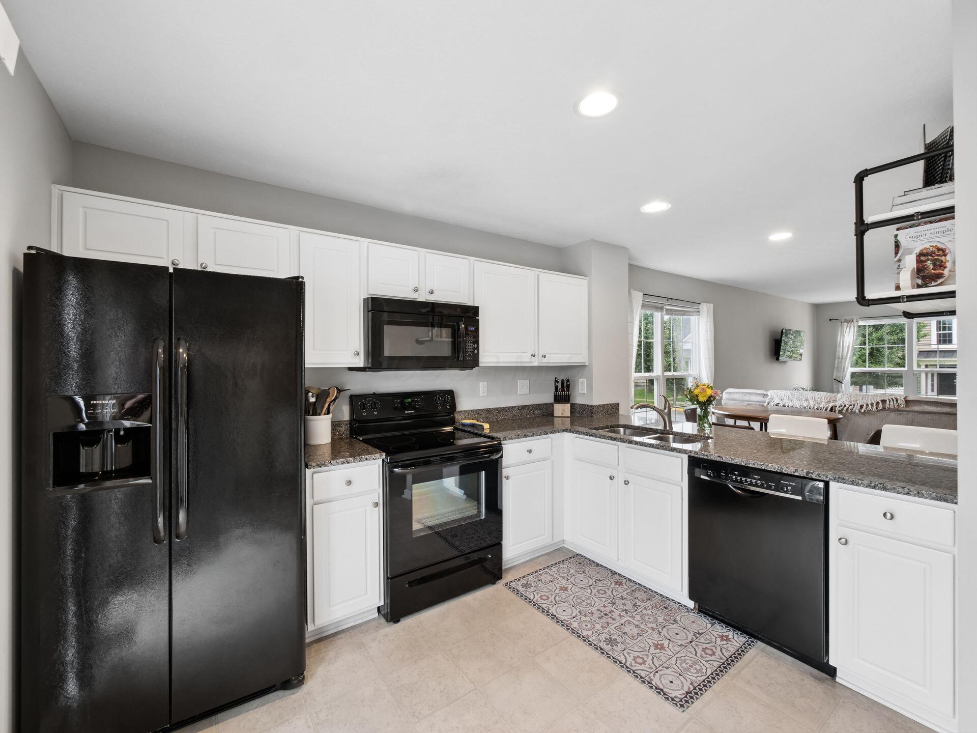 Kitchen with white cabinets, black appliances, granite counters, and a patterned runner opening to a dining and living area.