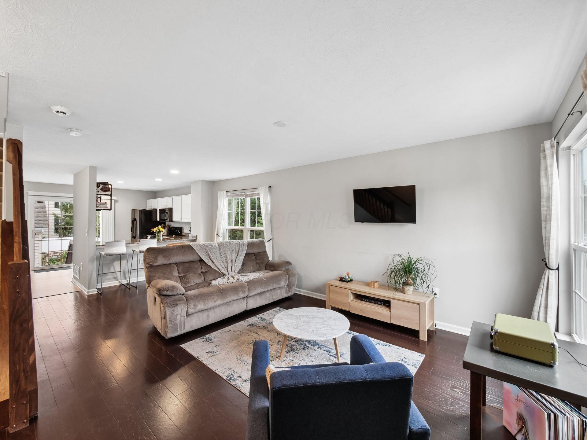 Open-plan living room with gray sofa, wall-mounted TV, large windows, and kitchen counter with stools.