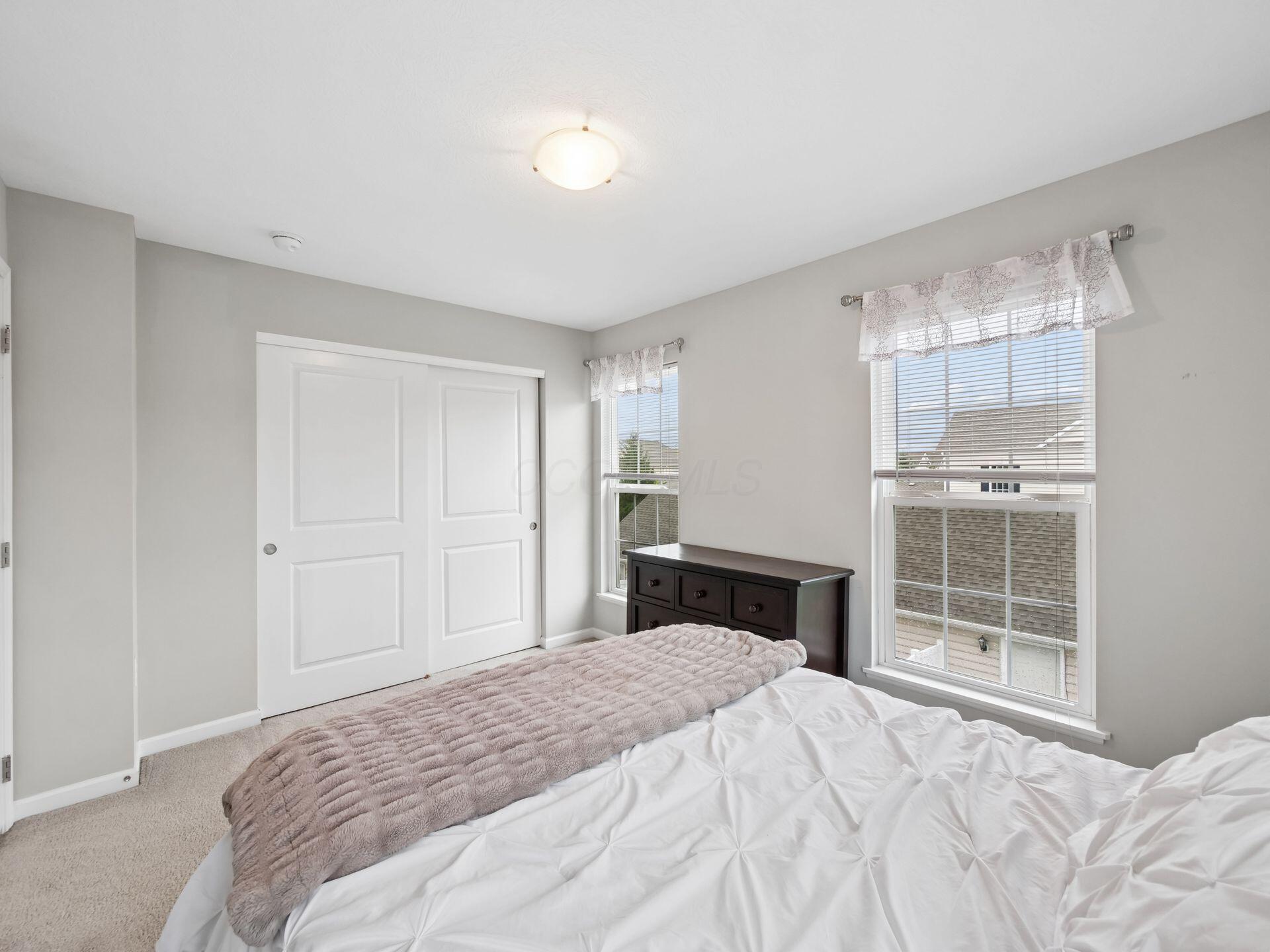 Bright bedroom with a white bed, taupe throw, dark dresser, and two windows.