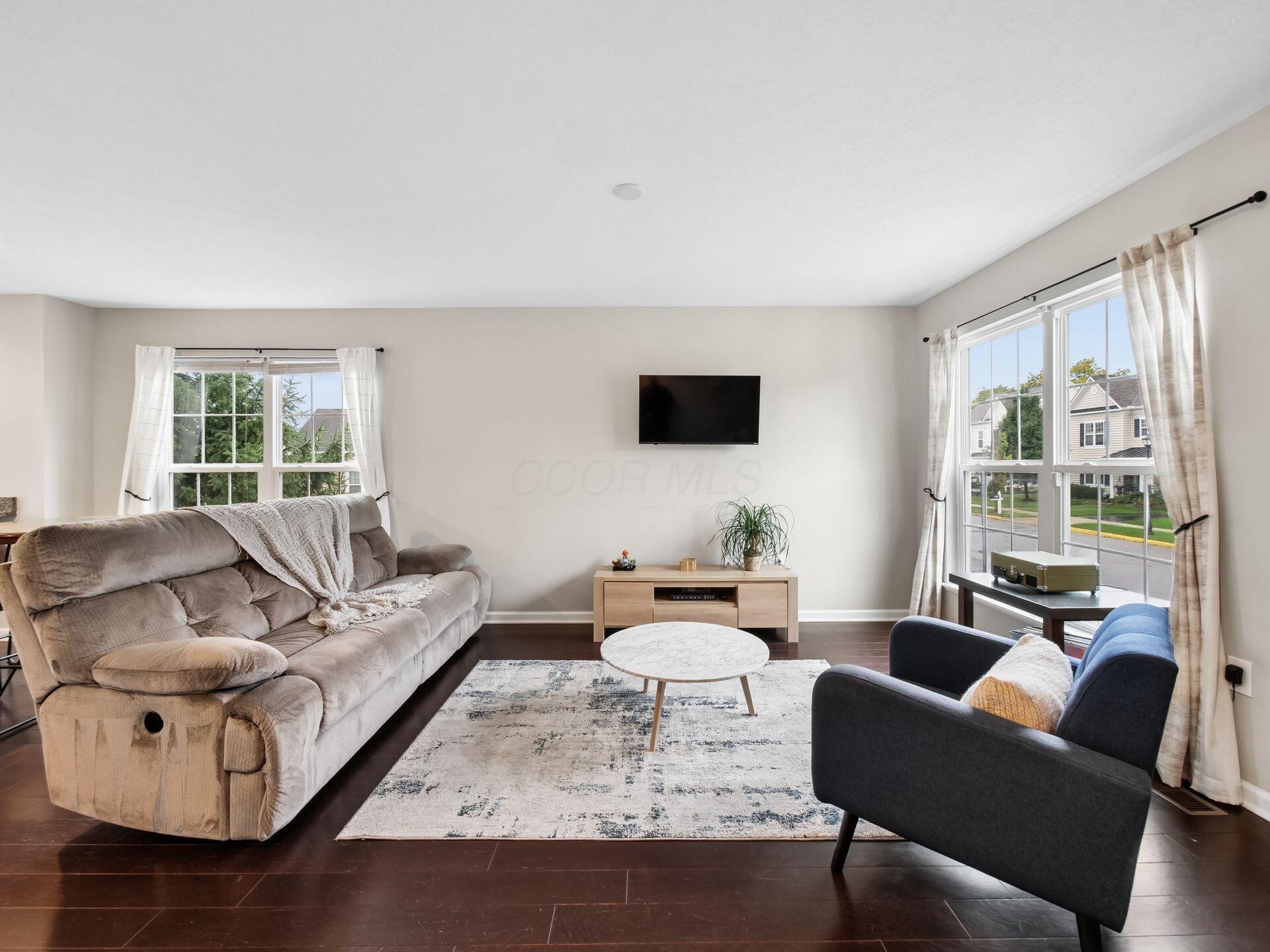 Living room with a beige reclining sofa, blue armchair, wall-mounted television, and large windows.