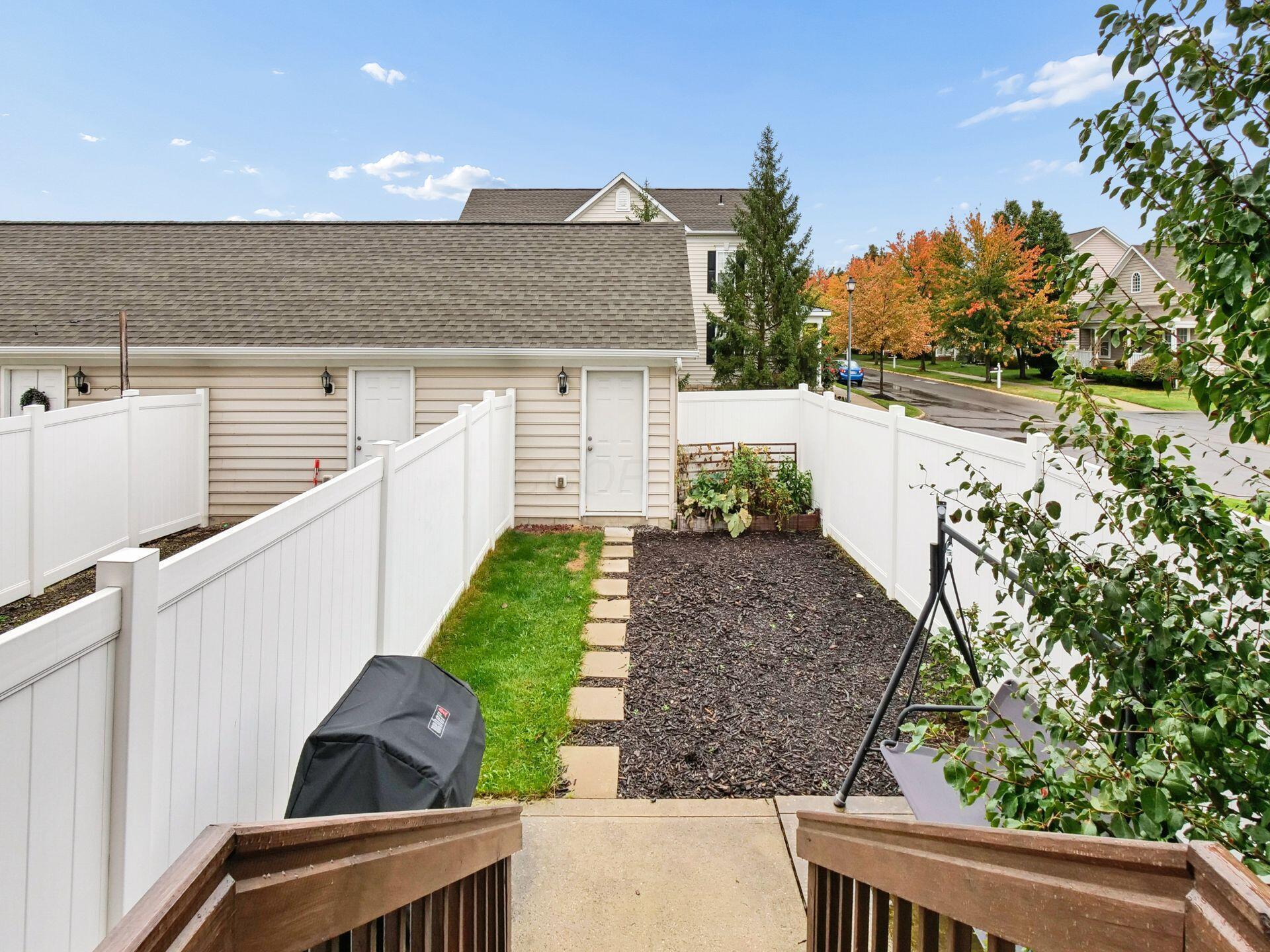 Fenced backyard with stepping stones, garden beds, a covered grill, and a hanging chair.