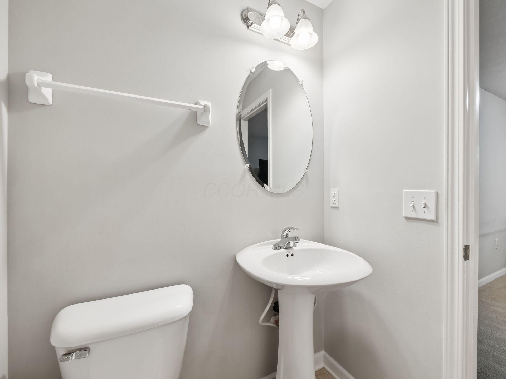 Bathroom with a white pedestal sink, oval mirror, and toilet beside an open doorway.