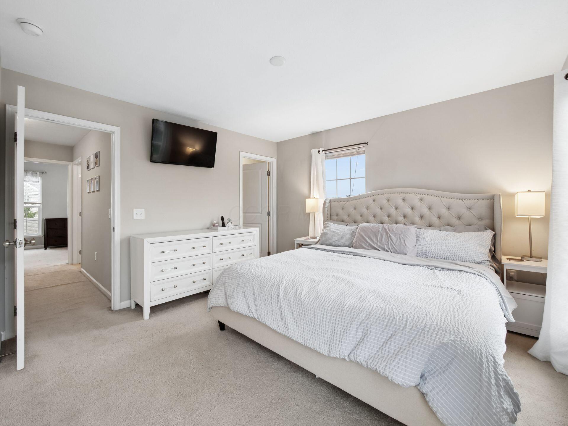 Furnished bedroom with a tufted headboard, white dresser, wall-mounted television, and curtained window.