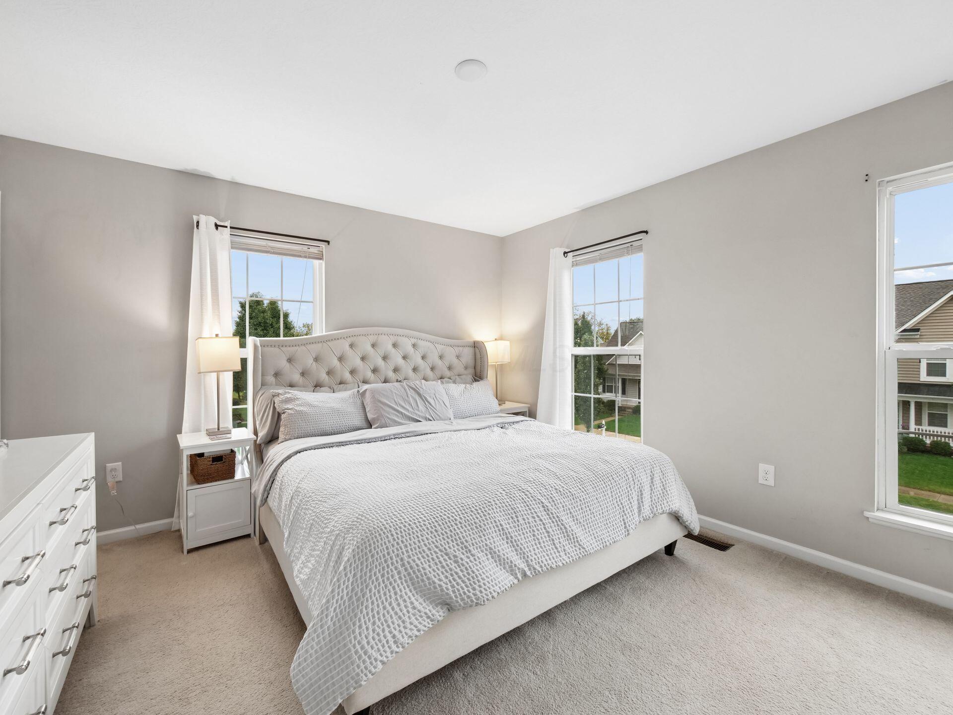 Staged bedroom with a tufted bed, white furnishings, and three windows.