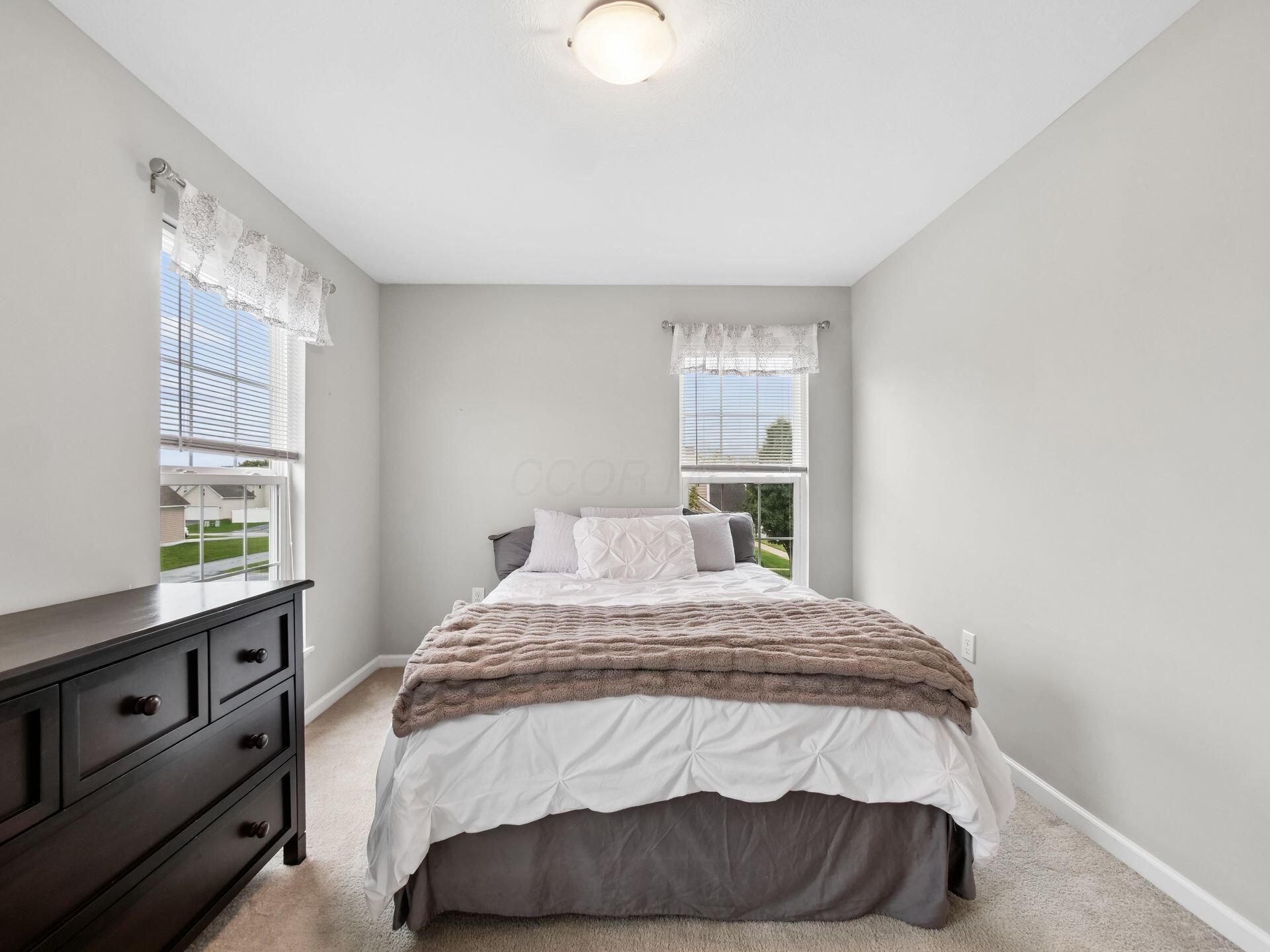 Furnished bedroom with a white bed, taupe blanket, two windows, and a dark wood dresser.