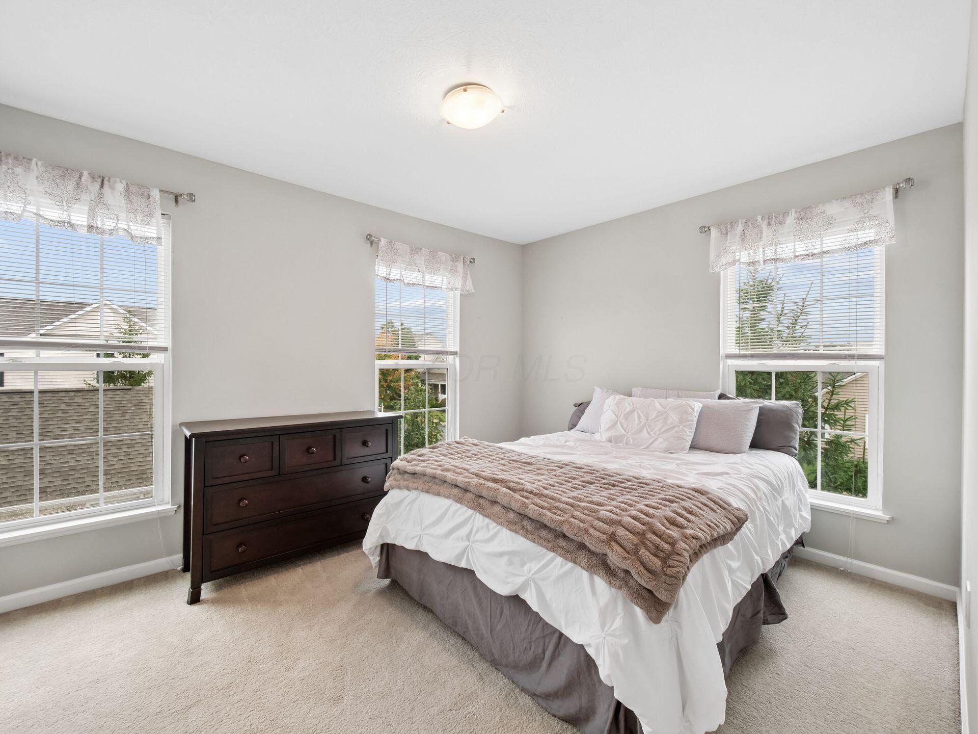 Bedroom with a white bed, taupe throw, dark wood dresser, and three windows.