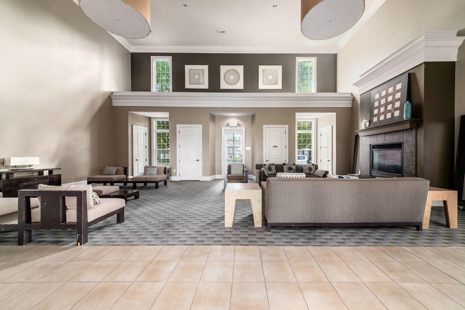 Furnished lounge with a double-height ceiling, tall windows, patterned carpet, and a large fireplace.