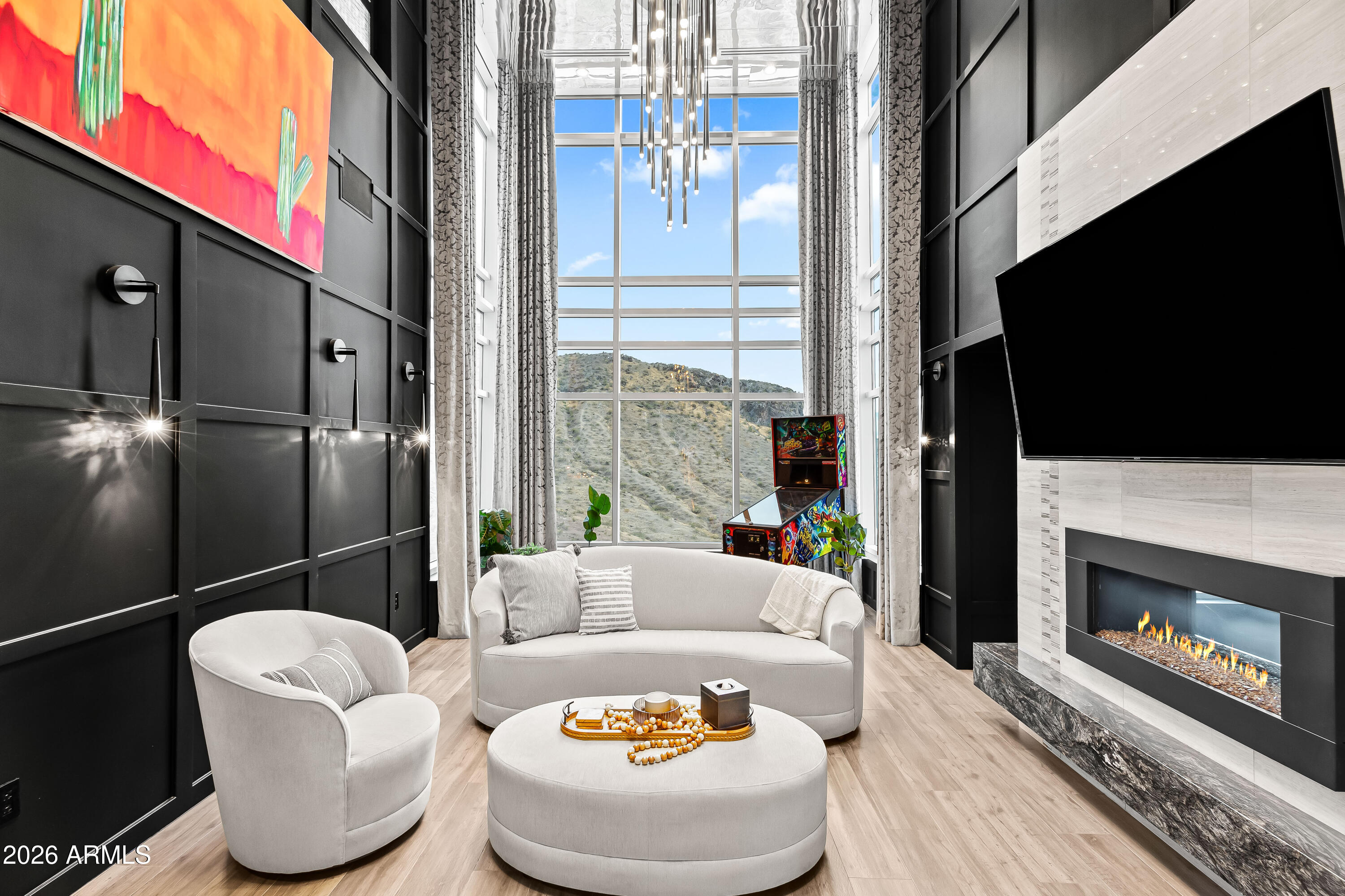 Living room with soaring windows, a fireplace, pale seating, and desert hills beyond.