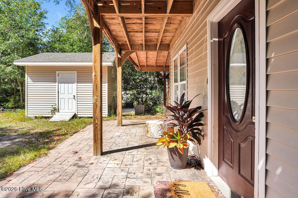 Covered paved patio beside a home, with timber supports, potted plants, and a detached outbuilding.