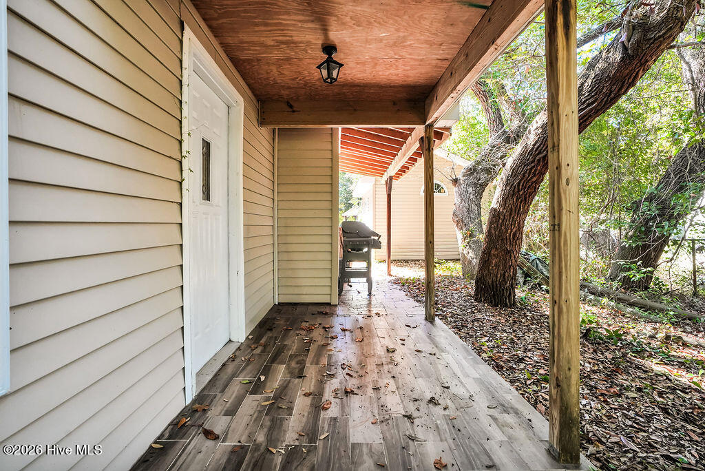 Covered side porch with weathered wood decking, beige siding, and mature trees alongside it.