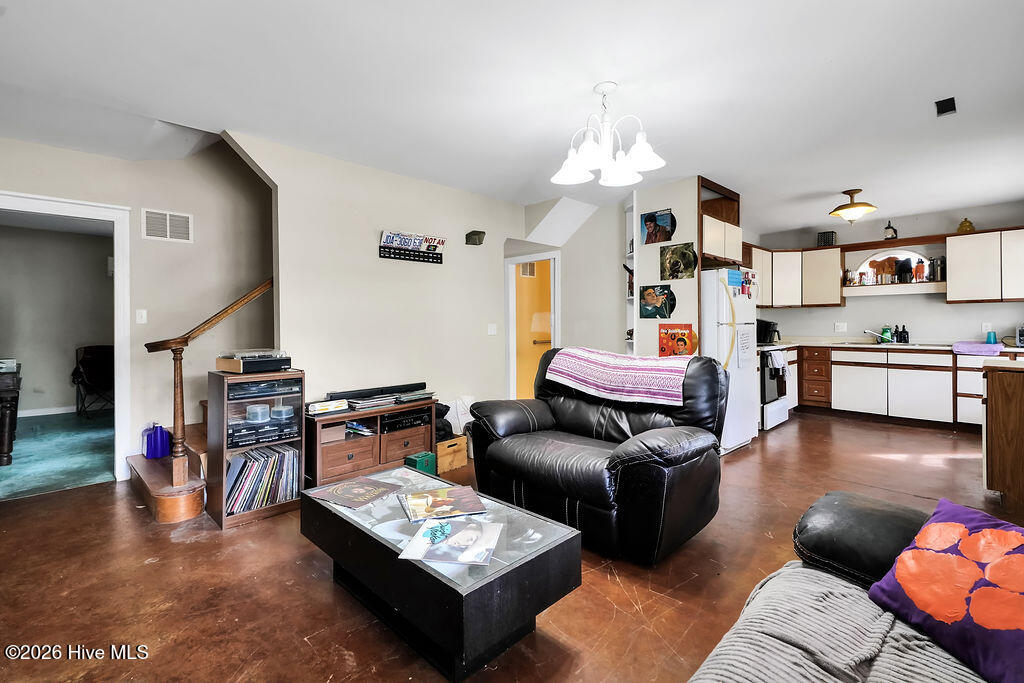 Open living room with a black leather sofa, brown flooring, and adjoining white-cabinet kitchen.