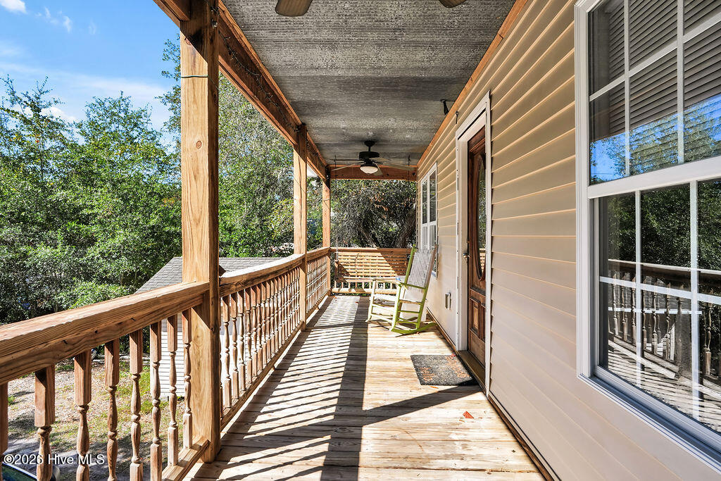Covered wooden porch with a ceiling fan, railing, and chairs beside tan siding and large windows.