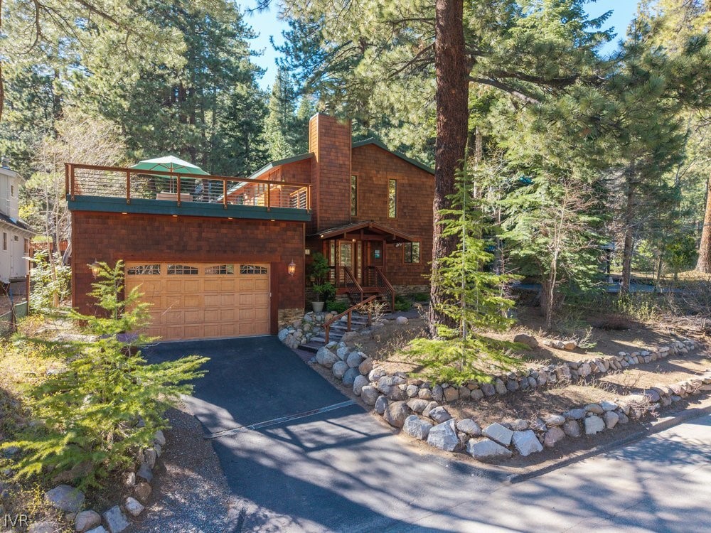 Wood-sided home among pine trees, with an elevated deck, two-car garage, and stone landscaping.