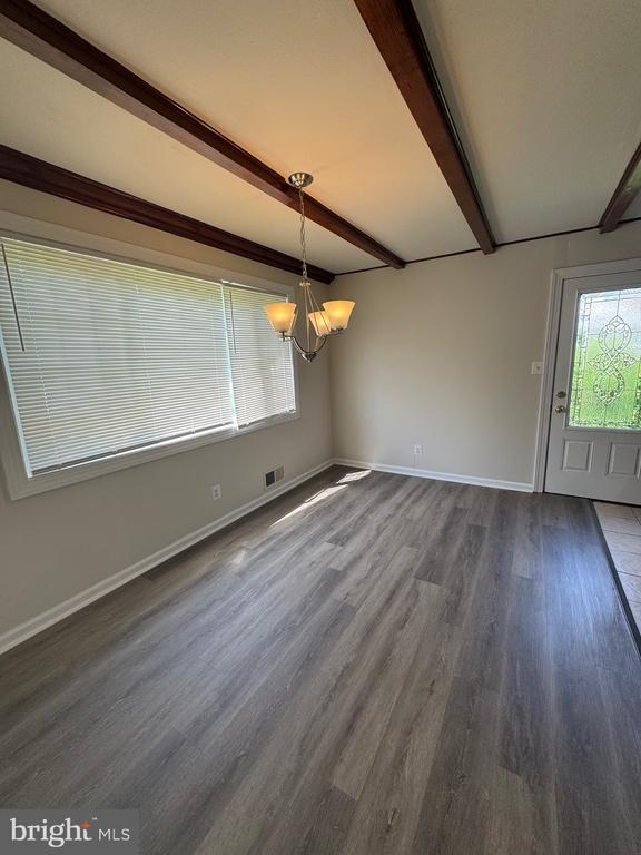 Empty dining room with exposed ceiling beams, chandelier, wide window, and gray plank flooring.
