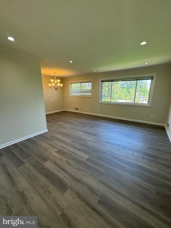 Vacant room with gray wood-look flooring, chandelier, recessed lights, and two windows overlooking greenery.