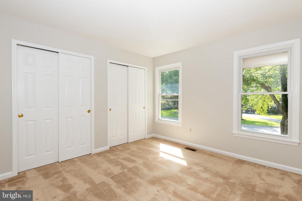 Empty bedroom with beige carpet, two closets, and windows overlooking leafy trees.