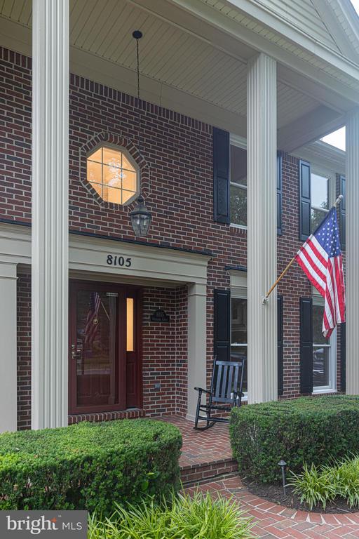 Red-brick home entrance with white columns, an American flag, and landscaped shrubs.