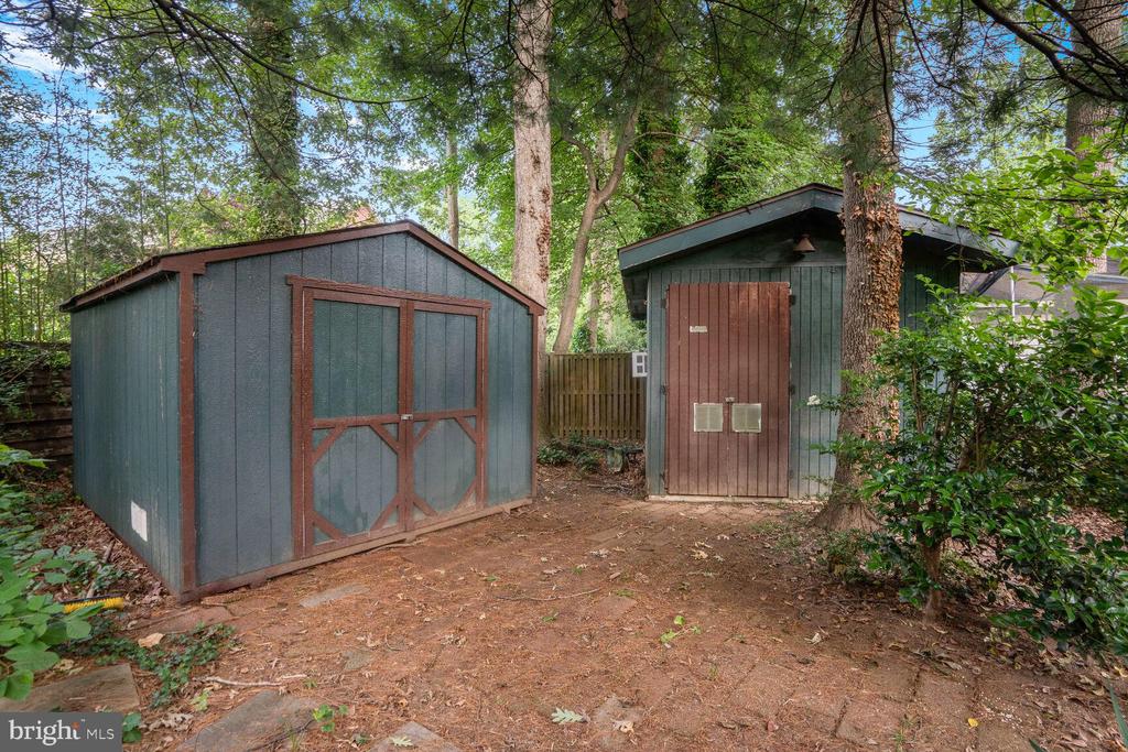 Two blue-green storage sheds with reddish-brown trim sit among trees in a wooded yard.