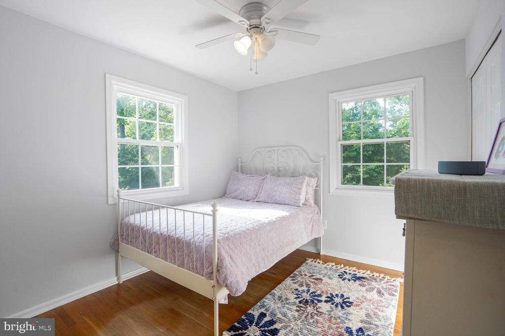 Bedroom with a white metal bed, pale pink bedding, two leafy-window views, and hardwood floors.