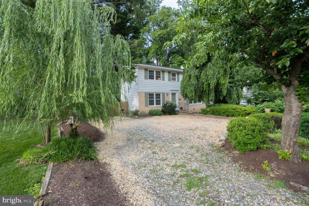 Two-story white-and-brick house behind a broad gravel driveway, framed by mature leafy trees.