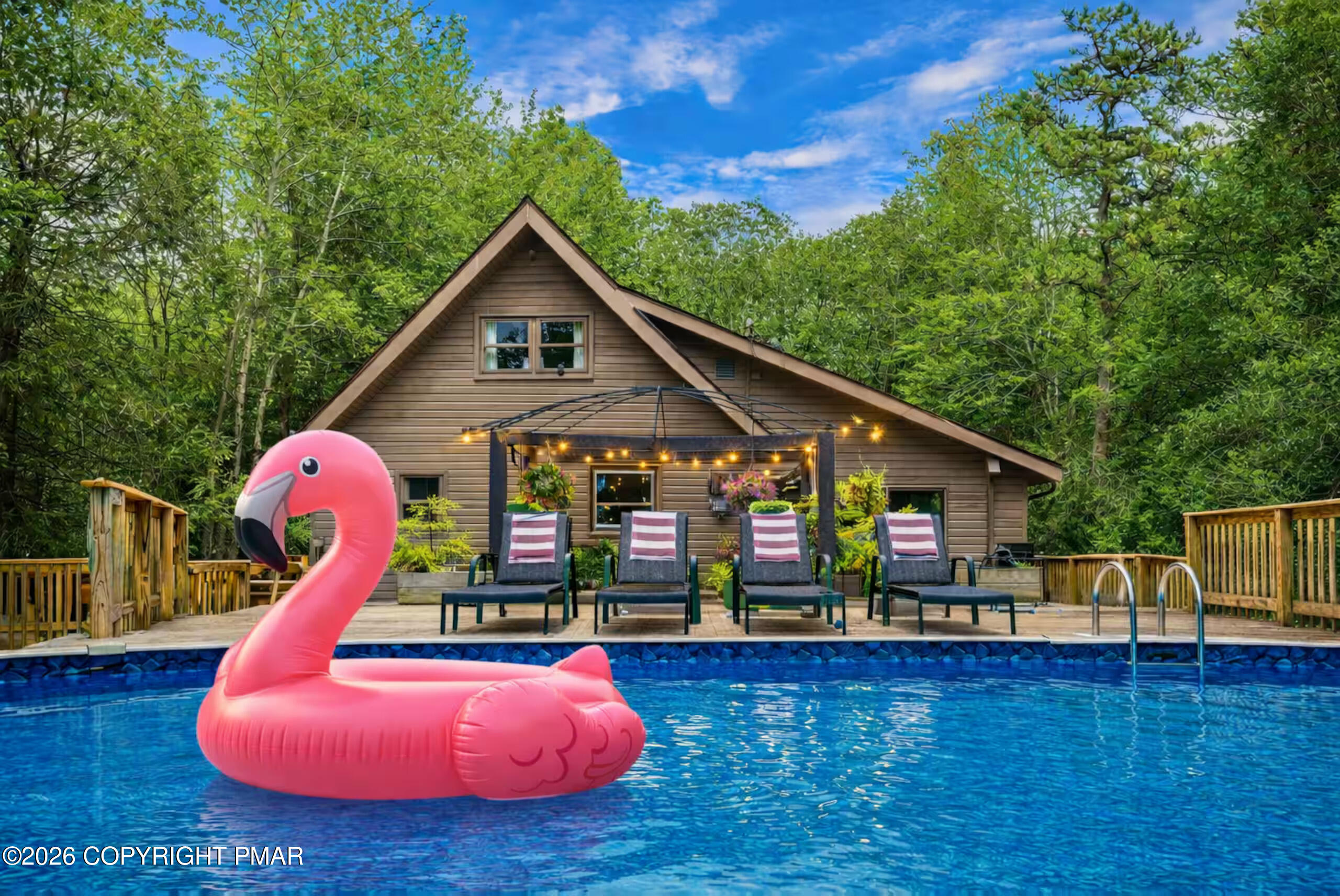 Wood-sided A-frame home behind a pool with a pink flamingo float and string-lit deck seating.
