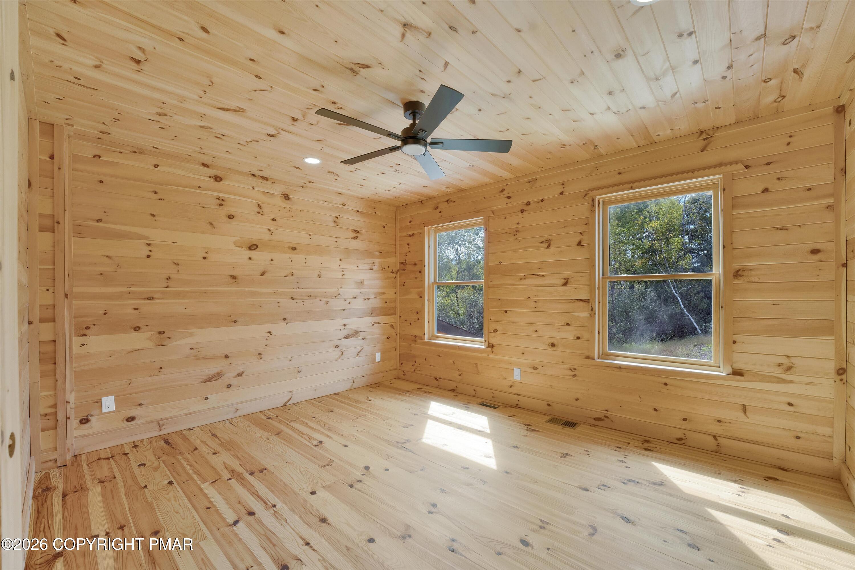 Empty pine-paneled room with matching wood floors, two windows, and a ceiling fan.