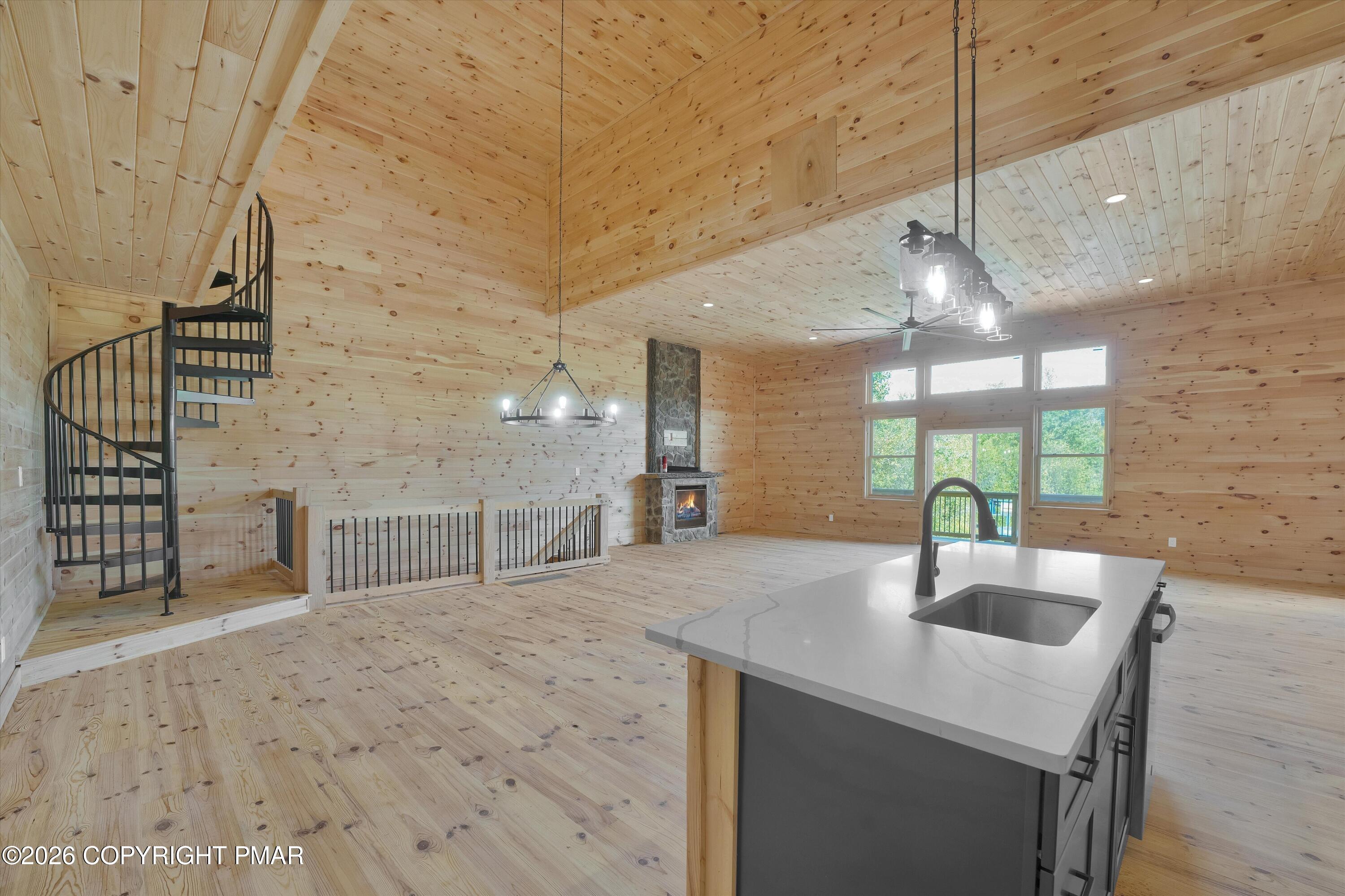 Vacant wood-paneled great room with a kitchen island, spiral staircase, stone fireplace, and vaulted ceiling.
