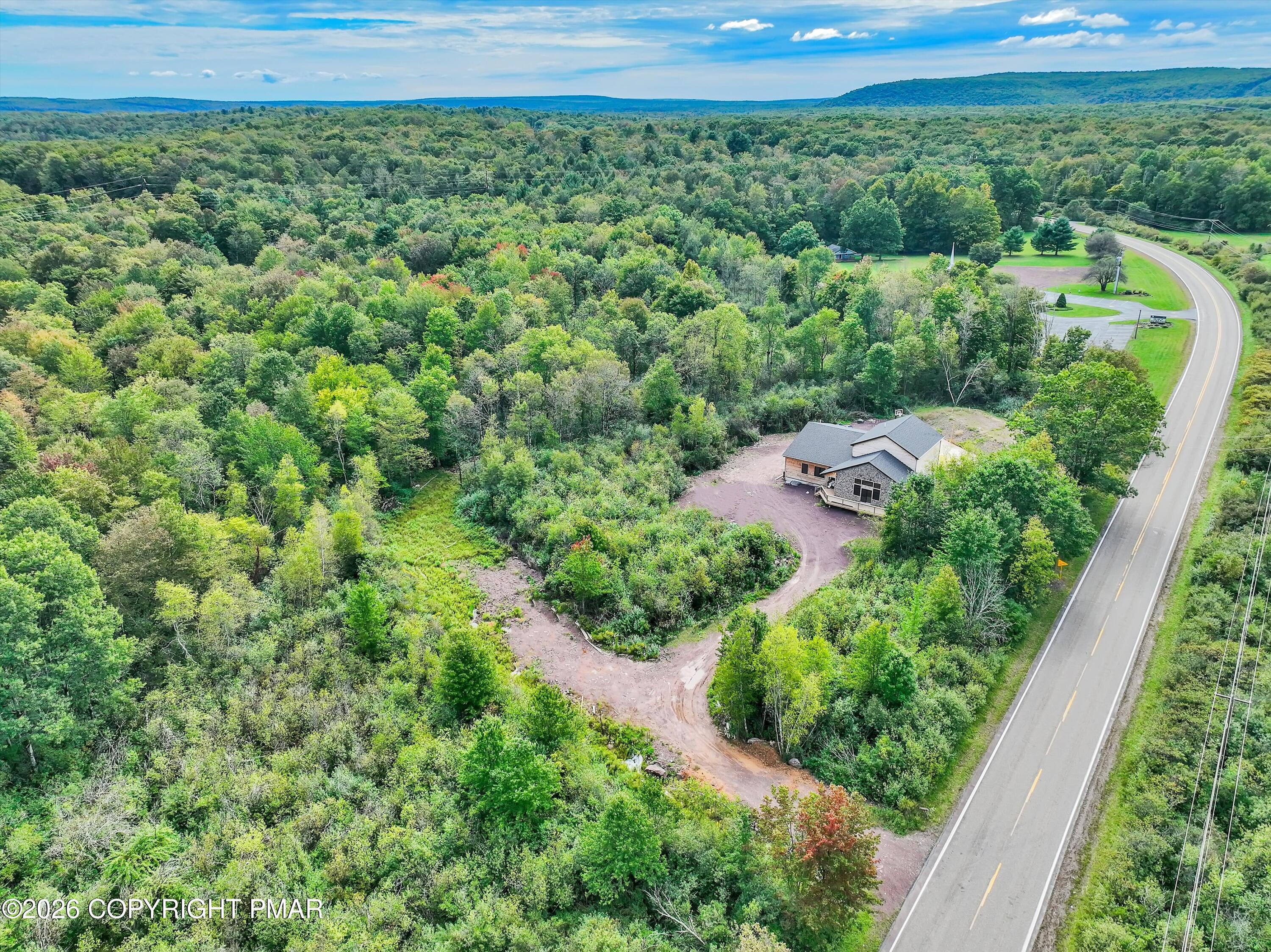 Aerial view of a house amid dense woods, beside a curving road and unfinished dirt driveway.