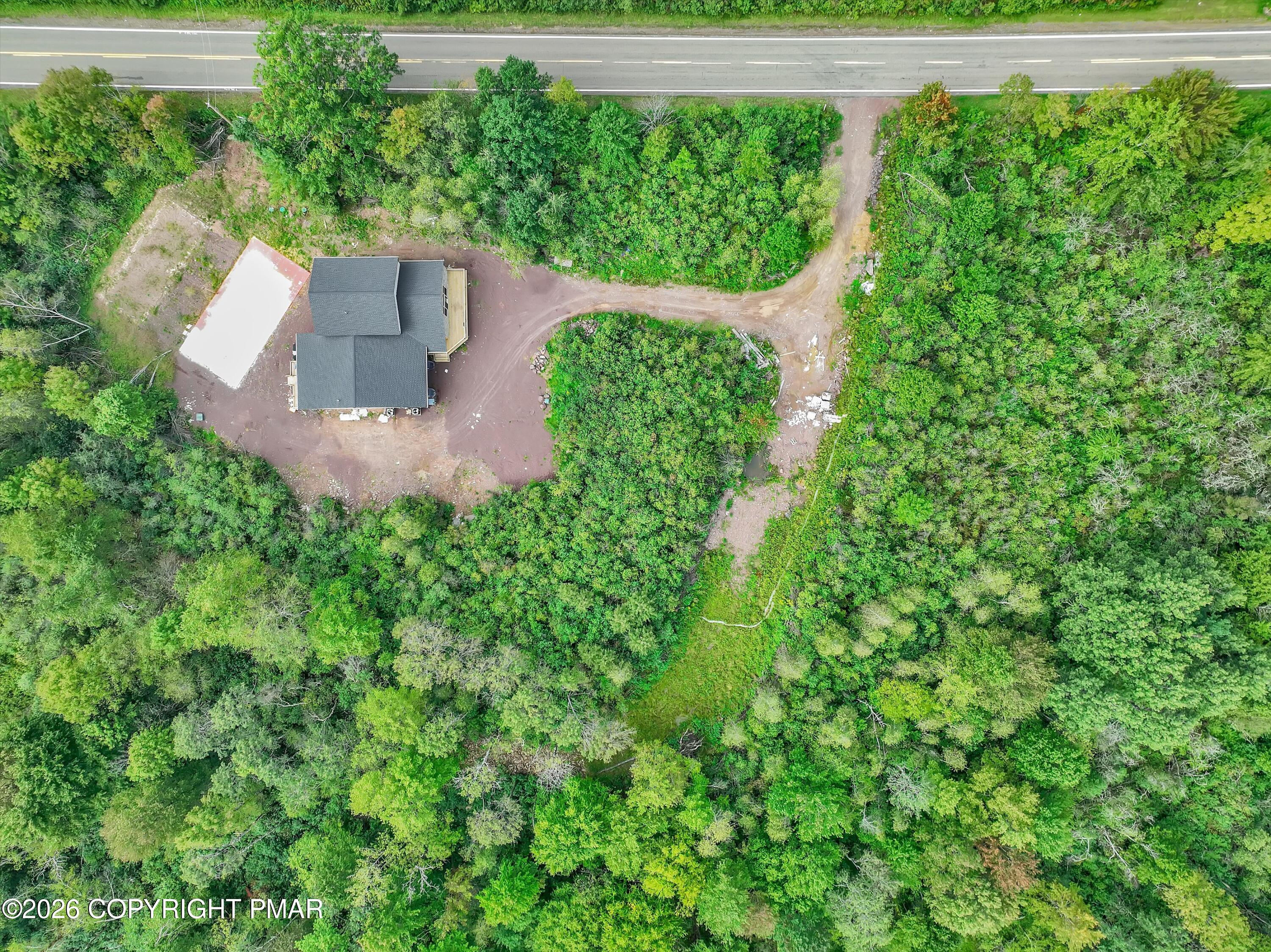 Aerial view of a house amid dense trees, with a curved dirt driveway and a separate white-roofed structure.
